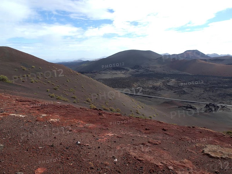 Volcanic landscape with red soil and hills under a slightly cloudy sky, barren landscape with roads, craters and small bushes, Lanzarote, spain