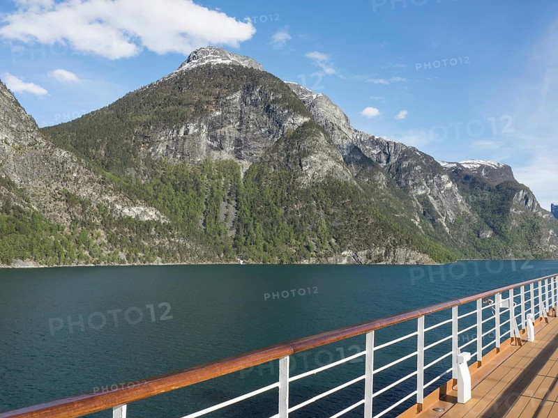 Ship deck in front of impressive mountain landscape with clear sea under blue sky with few clouds, ship railing in front of a panorama of mountains and sea, eidfjord, norway