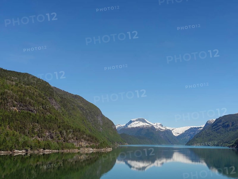 A calm lake, surrounded by snow-capped and forested mountains, under a clear blue sky, calm water in a fjord with snow-capped mountains and green slopes with trees, Eidjord, Norway, Europe