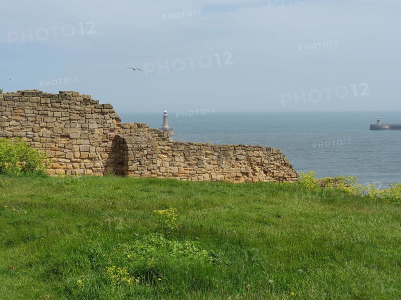 Remains of an old wall in front of a calm sea with a lighthouse in the distance, ruins and old stone walls by the sea, green fields with ruins, Newcastle, England, United Kingdom, Europe