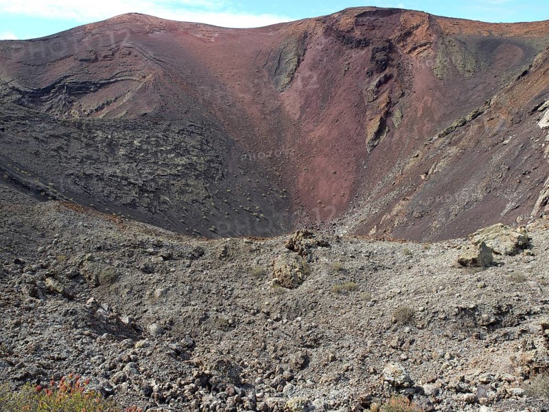 A broad volcano with a crater and red earth tones in a barren landscape, barren landscape with roads, craters and small bushes, Lanzarote, spain
