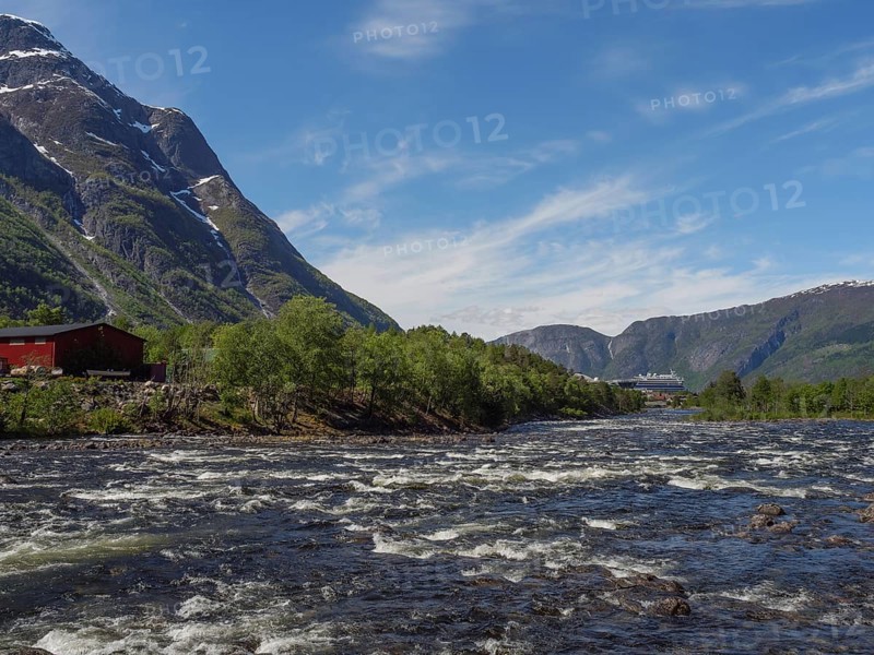 A turbulent river with mountains in the background, a few houses along the shore, wild river in a fjord with snow-capped mountains and green trees, eidfjord, Norway, Europe