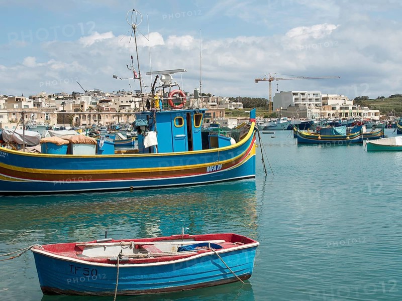 Harbour view with fishing boats and town in the background, a crane is visible, many colourful fishing boats in a harbour in the Mediterranean Sea, Marsaxlokk, malta