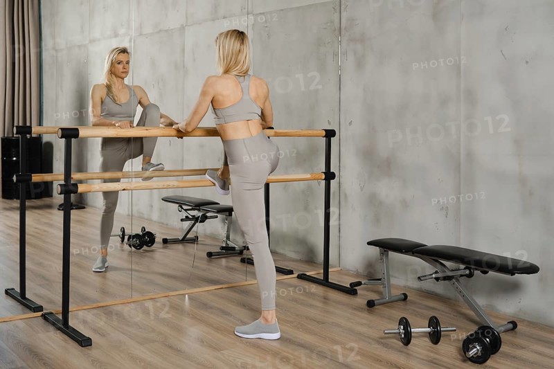 Active middle-aged woman does stretching exercises in gym using handrail. She is controlling her movements by looking at the wirror