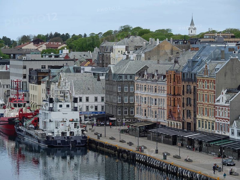 Waterfront of a town with many old and new buildings, yachts and fishing boats moored at the quay, old stone church and many gravestones at the waterfront with ships in the background, haugesund, norway