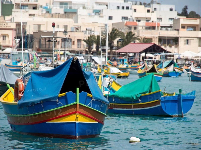 Colourful fishing boats in the harbour, partly covered with tarpaulins, in front of a coastal town in sunny weather, many colourful fishing boats in a harbour in the Mediterranean Sea, Marsaxlokk, malta