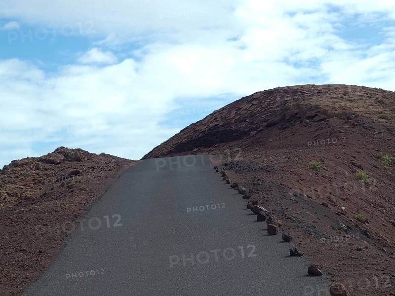 A tarmac road leads up a hill in a rocky volcanic landscape under a partly cloudy sky, barren landscape with roads, craters and small bushes, Lanzarote, spain