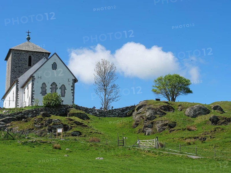 White church with stone tower on a rocky hill, tree and blue sky, old stone church and many gravestones by the water with ships in the background, haugesund, norway