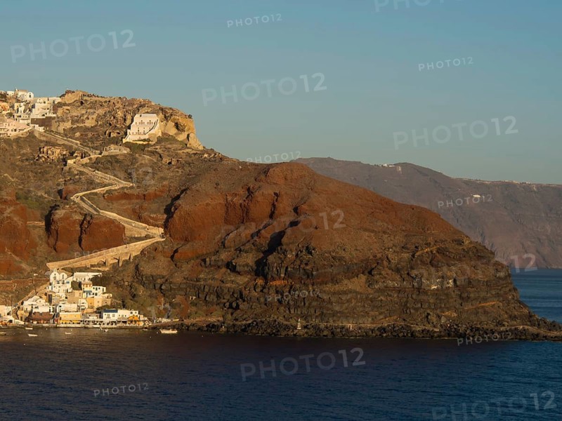 A coastline of Santorini with steep cliffs and houses rising above the azure blue sea, rocky island in the sea with white houses against a blue sky, Santorini, Greece, Europe