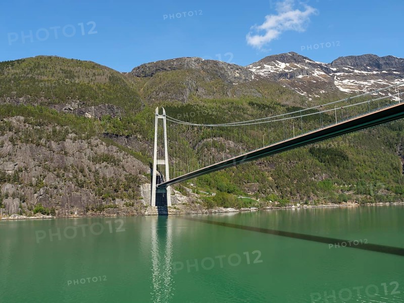 High bridge over a calm lake, surrounded by wooded and rocky hills under a bright blue sky, bridge in a fjord with snow-capped mountains and green trees, greenish shimmering water, eidfjord, Norway, Europe