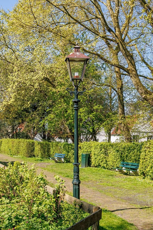 An antique lamppost stands in a well-kept park, surrounded by trees and green bushes on a sunny spring day, historic houses in a small village with small streets and trees in spring, Nes, Ameland, Netherlands