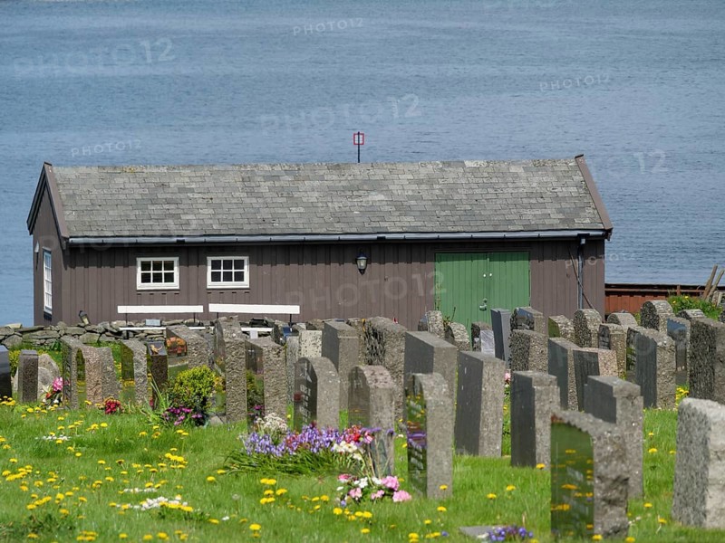 Wooden house behind a cemetery with many flowers on the shore of a lake, old stone church and many gravestones on the water with ships in the background, haugesund, norway