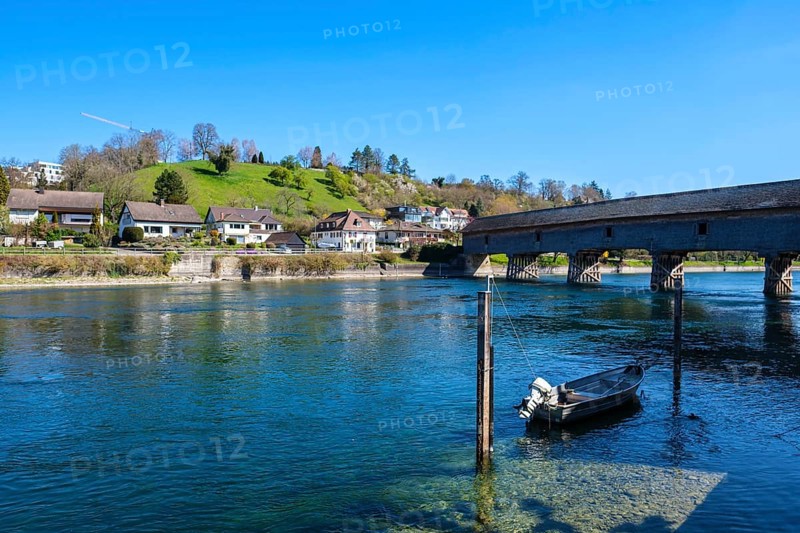 View of a calm river with a boat, a bridge and houses along the shore, embedded in a green hilly landscape, Diessenhofen, Thurgau, Switzerland, Europe