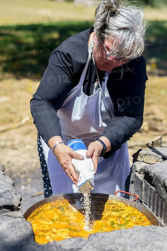 Woman cook in white apron pouring rice into a typical Spanish paella cooked in the countryside on the fire
