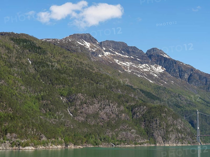 Massive mountains with partly forested slopes under a clear blue sky, some snow-capped peaks and a small waterfall, bridge in a fjord with snow-capped mountains and green trees, greenish shimmering water, eidfjord, Norway, Europe