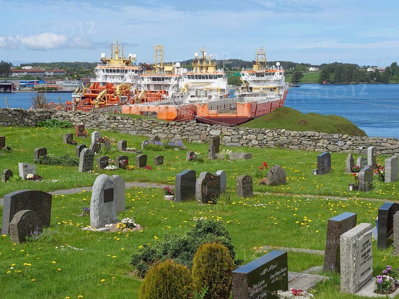 Cemetery with gravestones and three big orange ships by the sea, under a blue, slightly cloudy sky, old stone church and many gravestones by the water with ships in the background, haugesund, norway