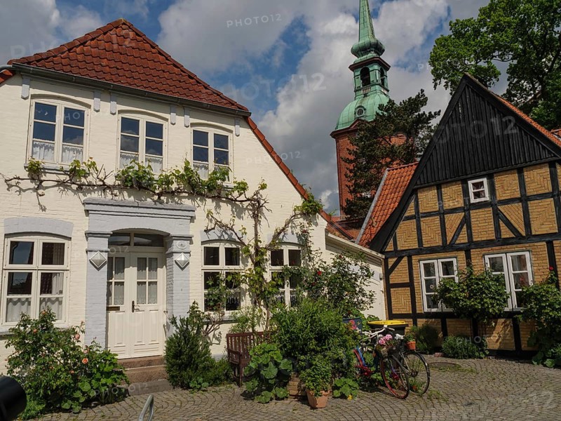 A white house with a red tiled roof next to a traditional half-timbered house under a blue sky with clouds, two houses, a church tower and bicycles decorated with flowers, kappeln, germany