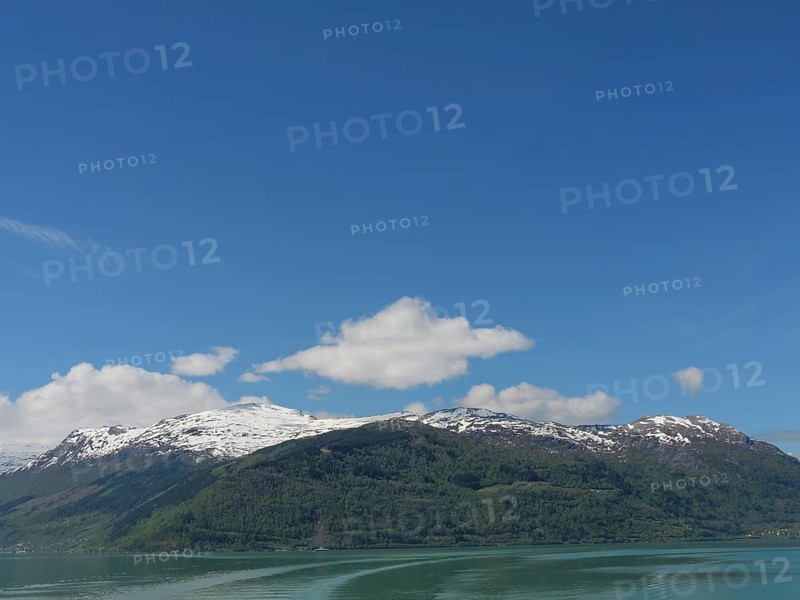 A mountain with snow peaks under a blue sky with clouds, in front of a calm lake, greenish shimmering water in a fjord with snow-covered mountains and green trees, eidfjord, Norway, Europe