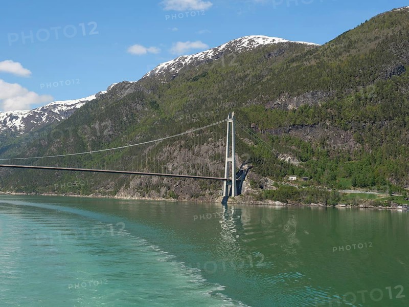 Vast landscape with a bridge stretching over a calm lake and snow-capped mountains under a blue sky, bridge in a fjord with snow-capped mountains and green trees, greenish shimmering water, eidfjord, Norway, Europe