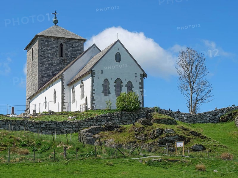 White church with stone tower on a rocky hill under blue sky with clouds, old stone church and many gravestones at the water with ships in the background, haugesund, norway