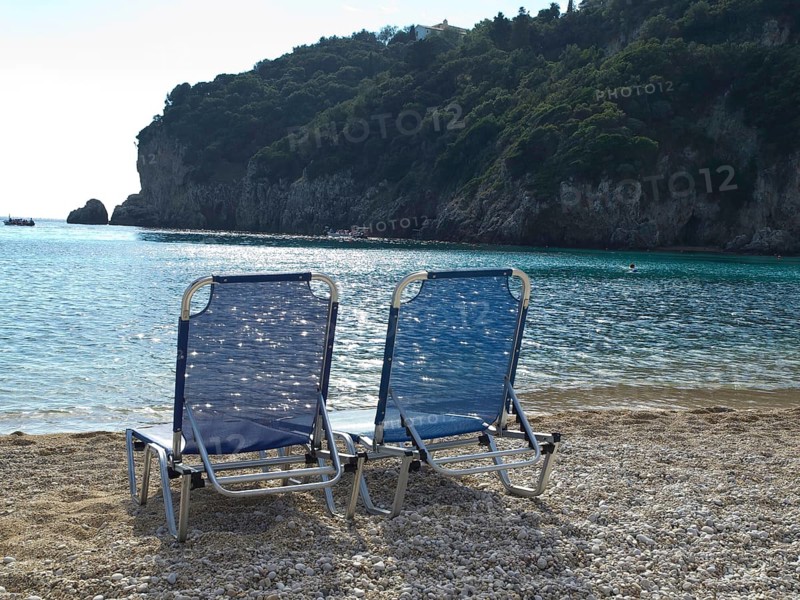 Two blue deckchairs on the beach with calm seascape and rocks in the background, umbrellas and sunbeds on a small beach with green blue water, corfu, greece