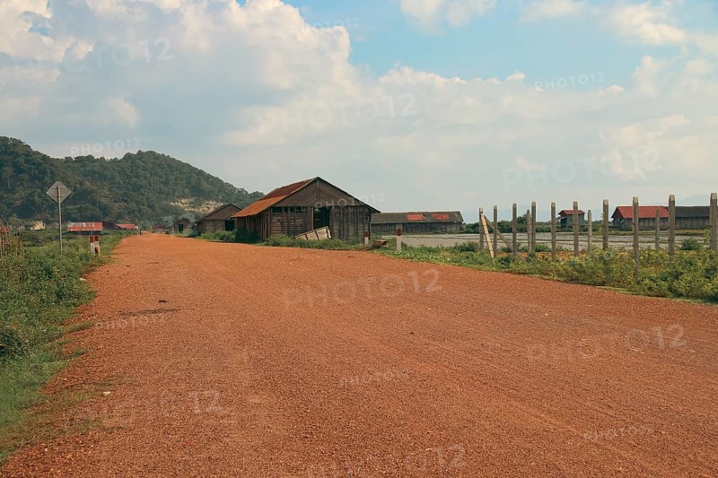 A serene rural landscape with a dirt road leading past wooden salt storage houses and mountains in the background. Fish Islad, Kampot Cambodia