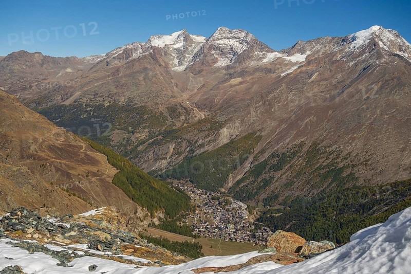 View down the valley to a village surrounded by snow covered mountains under a bright blue sky, snow on high mountains in the alps, blue sky over mountains, Saas fee, Switzerland, Europe