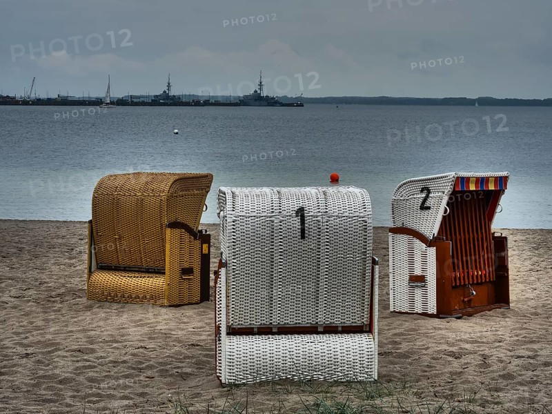 Three beach chairs stand on the sandy beach in front of a calm sea under a cloudy sky. Ships are visible on the horizon, beach chairs on a quiet sandy beach by the sea, clouds over the sea, Eckernfoerde, germany
