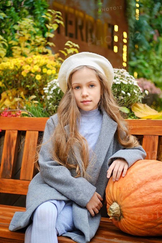 Cute little girl in a beret and woolen coat sits on bench leaning on pumpkin