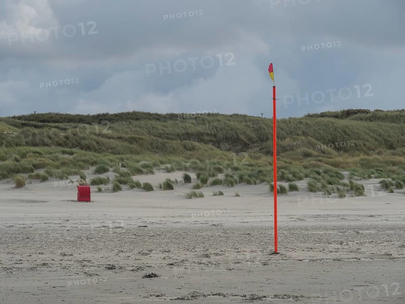 A lonely flagpole stands on a sandy beach in front of a hilly dune landscape, colourful beach chairs on the beach and in the dunes under an overcast sky, red wooden boat and yellow beach chair, Juist, Germany, Europe
