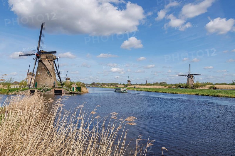 Windmills and a boat on a river under a blue sky with white clouds, many historic windmills on a river in the middle of fields, Kinderdijk, Netherlands