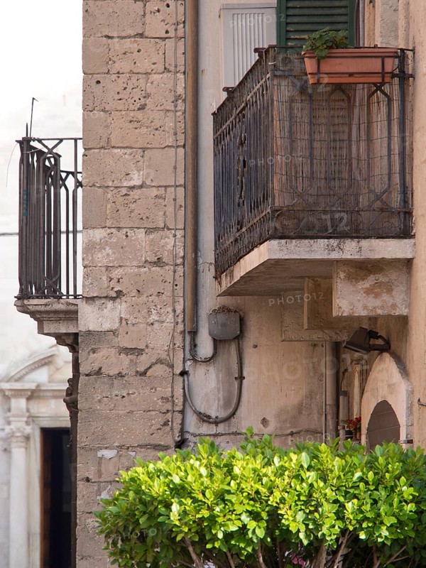 An old balcony on a stone facade with a flower pot and a wild green bush in the foreground, small balconies decorated with flowers on old houses, bari, italy