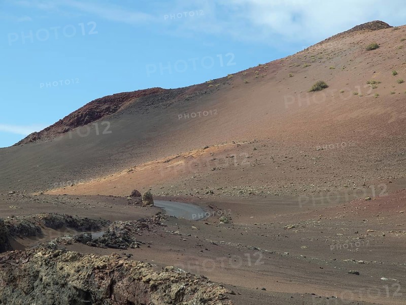 A rocky volcano with a narrow road, surrounded by lava and ash under a slightly cloudy sky, barren landscape with roads, craters and small bushes, Lanzarote, spain