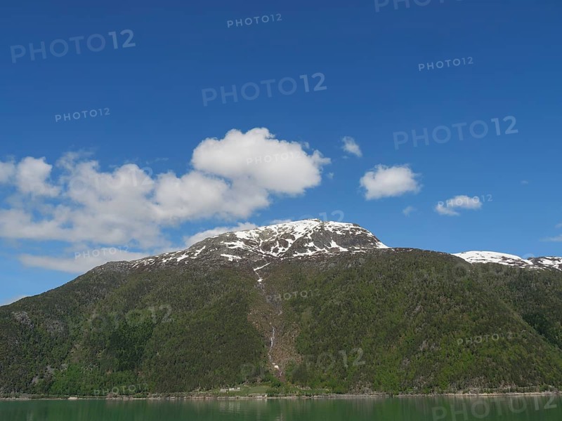 Wide view of snow-capped mountains under a wide blue sky, greenish shimmering water in a fjord with snow-capped mountains and green trees, eidfjord, Norway, Europe