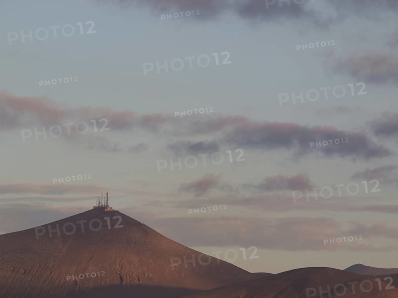 Mountain with antennas on the summit, surrounded by clouds in the soft light of dusk, barren landscape with roads, craters and small bushes, Lanzarote, spain
