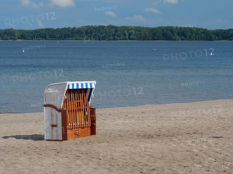 An empty beach chair stands on the sandy beach of a lake under a blue sky with some clouds, lonely beach chair on a quiet beach with a tree in the background, Eckernfoerde, germany
