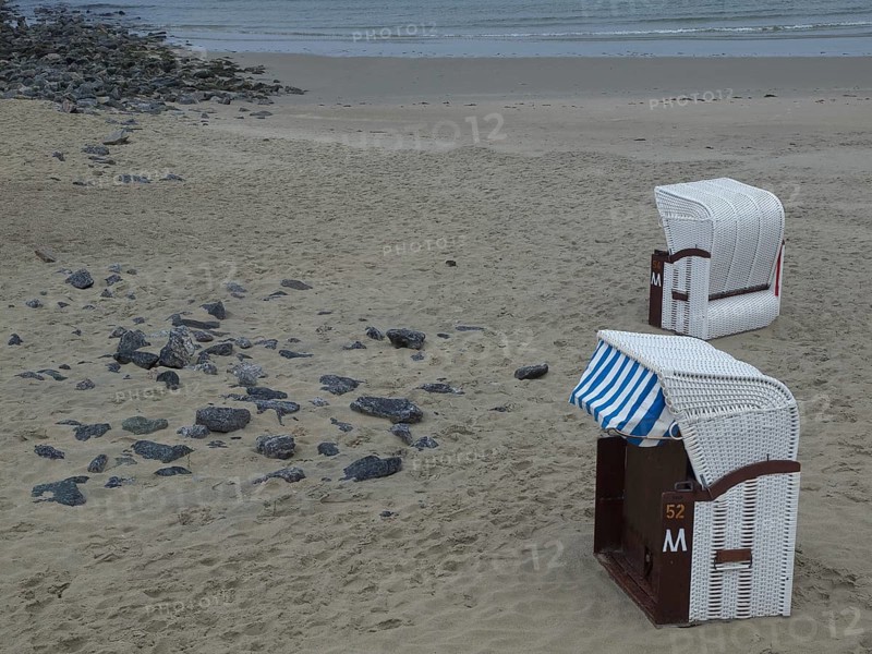 Two white beach chairs on an empty beach, some stones in the sand and the calm sea in the background, beach chairs and beach tents by the sea under a cloudy sky, boardwalk in the sand, Borkum, germany