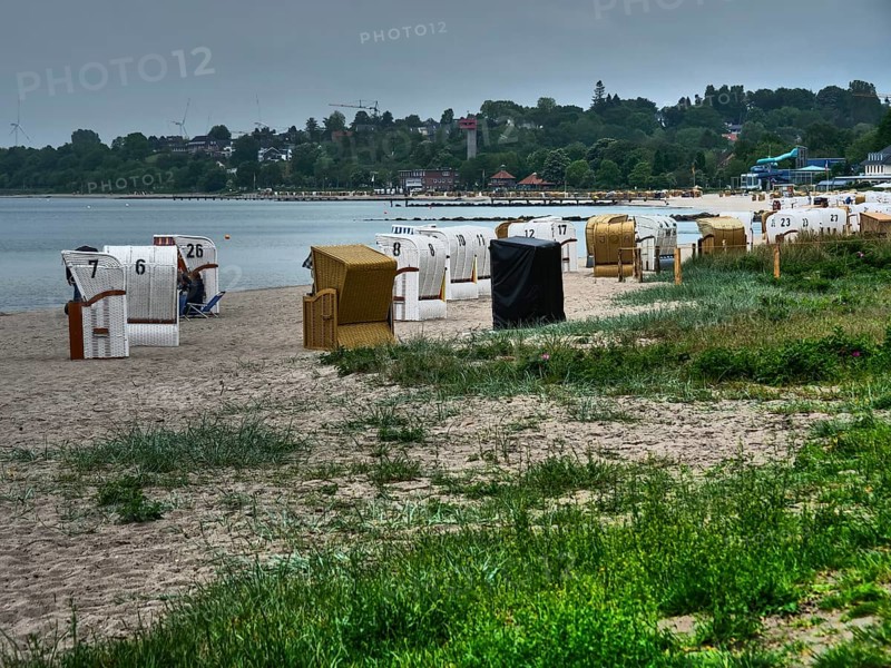Several beach chairs stand on a sandy beach along the coast with dense vegetation and hills in the background. Clouds cover the sky, beach chairs on a quiet sandy beach by the sea, clouds over the sea, Eckernfoerde, germany