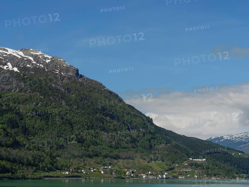 Small settlement in front of a green mountainside and snow-capped peaks under a clear sky, greenish shimmering water in a fjord with snow-capped mountains and green trees, eidfjord, Norway, Europe