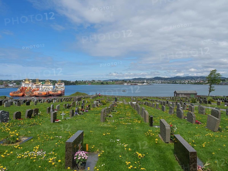 Cemetery with many gravestones near a body of water with ships under a blue, slightly cloudy sky, old stone church and many gravestones by the water with ships in the background, haugesund, norway