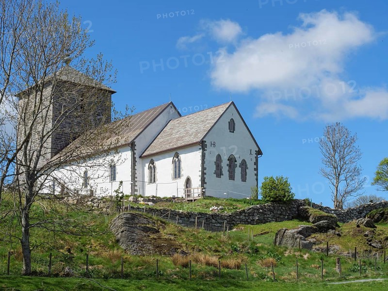 White church with stone tower on a hill under a blue sky, trees in the foreground, old stone church and many gravestones on the water with ships in the background, haugesund, norway