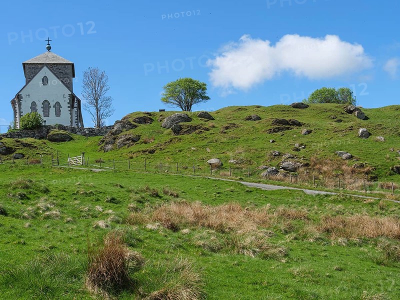Church with stone tower on hilly landscape under blue sky with clouds, trees in the background, old stone church and many gravestones at the water with ships in the background, haugesund, norway