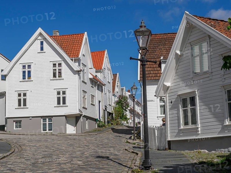White wooden houses with red tiled roofs along a cobblestone street, street lamp, bright blue sky, white wooden houses with red roofs against a blue sky, trees and white fences along the street, stavanger, norway