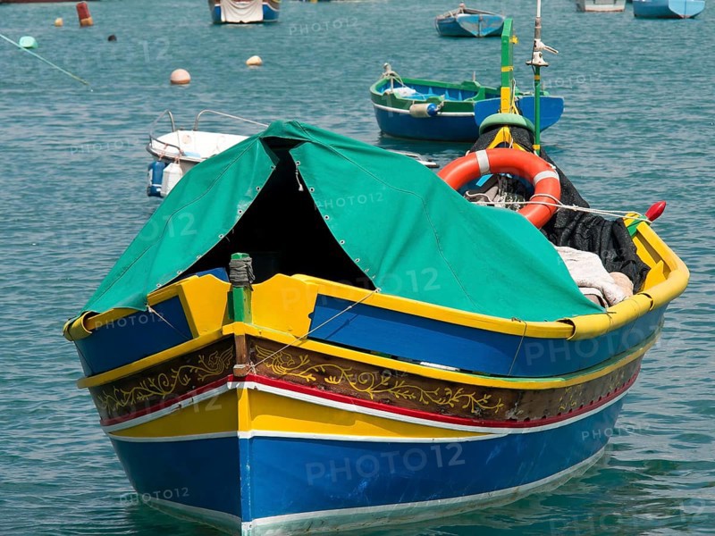 A colourful fishing boat with green fabric cover and lifebuoy at anchor, many colourful fishing boats in a harbour in the Mediterranean, Marsaxlokk, malta