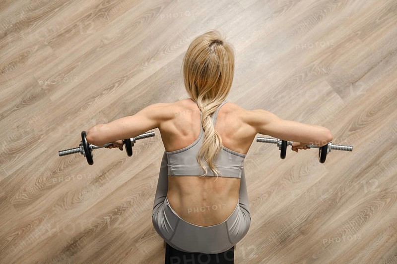 Top view of female bodybuilder pushing dumbbell to the sides while sitting on bench in gym