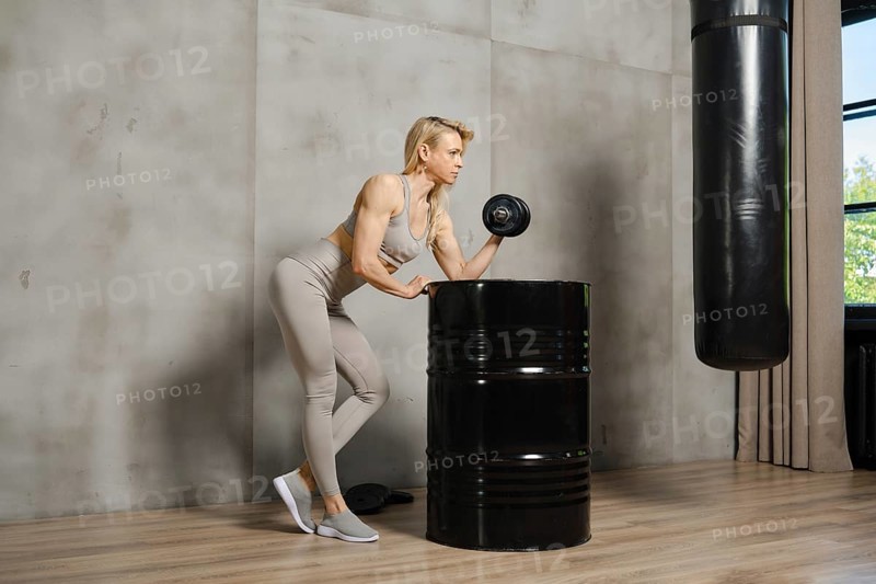 Female bodybuilder pushing dumbbell in the gym leaning on metal barrel