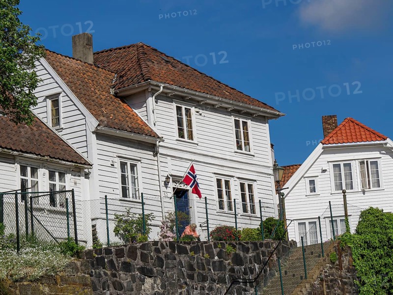 White wooden houses with red tiled roofs and norwegian flag in the foreground of a sunny day, white wooden houses with red roofs against a blue sky, trees and white fences along the road, stavanger, norway
