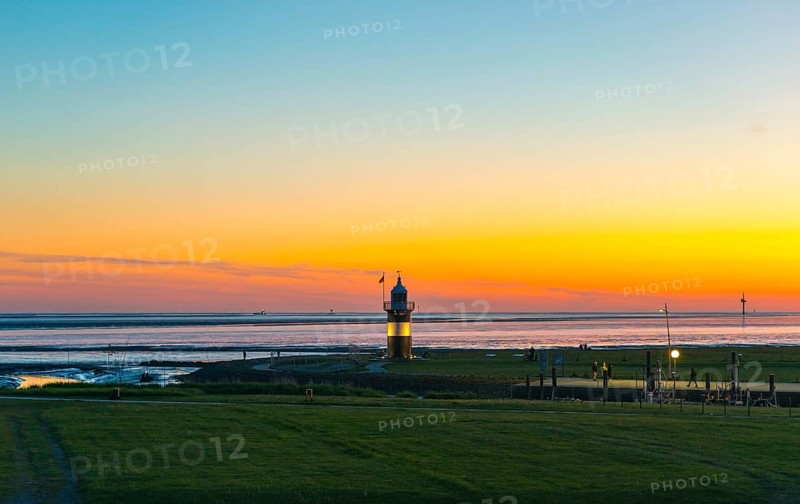 Black and white lighthouse, called 'Kleiner Preusse', stands on the coast of the North Sea under a colourful sky shortly after sunset and is illuminated, green meadow, sea, some people are walking, a tideway connects the cutter harbour Wremen with the Outer Weser and the sea, North Sea resort Wremen, Lower Saxony Wadden Sea National Park, World Heritage Site, Wurster North Sea coast, Land Wursten, district of Cuxhaven, Lower Saxony, Germany, Europe