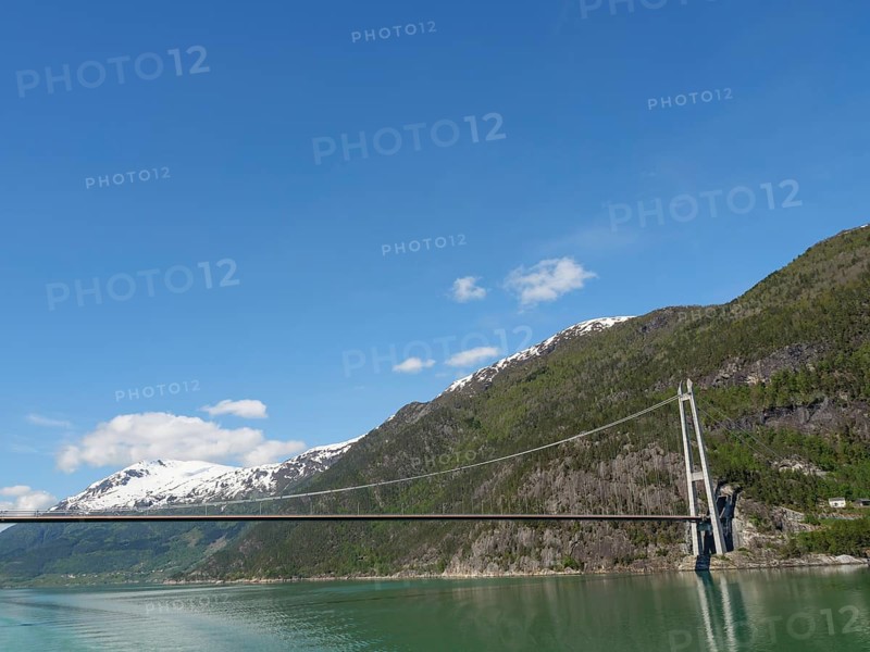 Wide view of a bridge and snow-covered mountains over a green lake and clear blue sky, bridge in a fjord with snow-covered mountains and green trees, greenish shimmering water, eidfjord, Norway, Europe