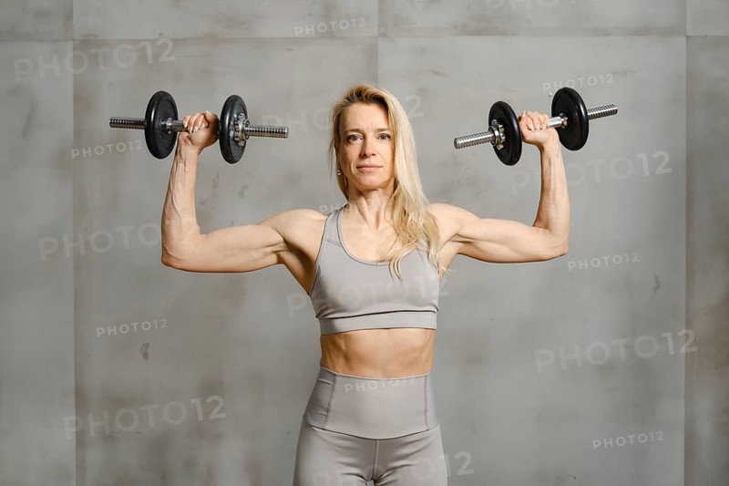A middle-aged woman stands near the grey wall of the gym and pushing dumbbells up